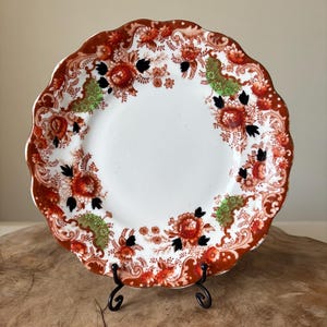May include: A white ceramic plate with a scalloped edge, featuring a floral pattern in shades of red, orange, black, and green. The plate is displayed on a black metal stand, showcasing its intricate design and vintage aesthetic.