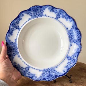 May include: A white ceramic bowl with a scalloped edge and a blue floral design. The rim is trimmed with gold. The bowl is held by a person's hand.
