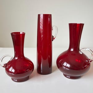 Puede incluir: Tres jarrones de cristal rojo rubí con asas de cristal transparente. Los jarrones varían en forma y tamaño, uno alto y cilíndrico y dos con cuerpos redondeados. El cristal tiene un acabado brillante que refleja la luz. Los jarrones se exhiben sobre una superficie blanca.