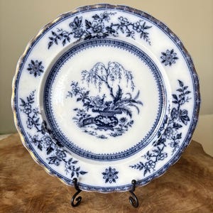 May include: White and blue decorative plate with a floral and tree design. The plate has a gold trim and is supported by a black metal stand. The plate's design includes flowers, butterflies, and a weeping willow tree.
