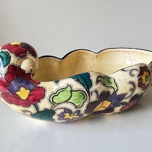 May include: A cream-colored ceramic bowl with a scalloped rim and a curled handle. The bowl is decorated with colorful floral patterns in red, purple, yellow, and green. The interior is a light beige color, and the rim is outlined in black.