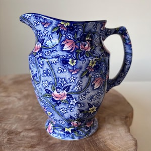 May include: Blue ceramic pitcher with a floral pattern. The pitcher features pink roses, yellow accents, and white flowers on a blue background. It has a handle and a spout for pouring.