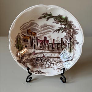 以下が含まれることがあります： 茶色と白の転写デザインが施された、スカラップエッジの装飾用プレート。 馬車のあるお城のシーンが描かれています。 このプレートは黒い金属製のスタンドに飾られており、そのヴィンテージ感を際立たせています。