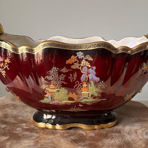 Op de afbeelding: Een bordeauxrode keramische plantenbak met gouden rand en handvatten. De plantenbak heeft een geschulpte rand en een zwarte voet met gouden accenten. De buitenkant is versierd met een kleurrijke, Aziatisch geïnspireerde landschapsscène.