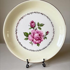 May include: A white ceramic plate with a gold rim and a pink rose design. The plate is on a black metal stand.