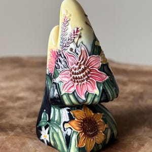 Peut inclure: Une pièce décorative en céramique avec un design empilé, avec un dessus jaune et une base sombre. Elle est ornée de motifs floraux détaillés, notamment des tournesols roses et oranges, et des feuilles vertes. Le design combine des éléments artistiques et naturels.