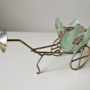 May include: A decorative, vintage-style cart with a light green glass bowl and metal wheels. The bowl is adorned with gold-colored leaf accents. The cart has a silver-colored cone at the front, and the overall design is reminiscent of a miniature carriage.