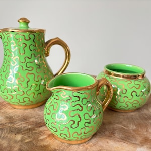 Może przedstawiać: Trzyczęściowy zestaw ceramiczny w żywym zielonym kolorze ze złotymi, kręconymi wzorami. Zestaw zawiera dzbanek z pokrywką, mały dzbanuszek do śmietanki i cukiernicę. Obręcze i uchwyty są złote.