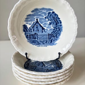 May include: A stack of white ceramic bowls with a blue and white cottage scene. The bowls have a scalloped edge and are stacked on a black metal stand. The text "A Cottage in a Cornfield" is visible.