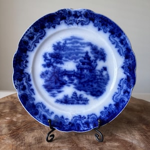 Puede incluir: Un plato redondo antiguo azul y blanco con borde festoneado. El plato presenta una escena detallada de paisaje con una pagoda y árboles. El borde tiene un patrón floral decorativo. El plato se exhibe sobre un soporte de metal negro.