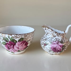 Könnte beinhalten: Ein weißes Porzellan-Zucker- und Sahne-Set. Das Set hat ein Blumendesign mit rosa Rosen, grünen Blättern und goldenen Akzenten. Der Rand jedes Stücks ist mit Gold verziert.