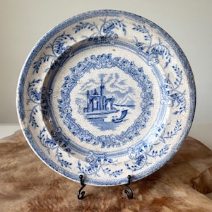 May include: Antique blue and white ceramic plate with a detailed landscape scene in the center. The plate features a decorative floral border and a slightly aged appearance, suggesting its vintage origin. The plate is displayed on a black metal stand.
