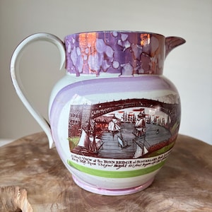 Puede incluir: Jarra de cerámica vintage con asa y boquilla blancas. La jarra presenta un diseño morado y blanco con una ilustración detallada de un puente y barcos. El texto en la jarra dice "A West View of the IRON BRIDGE at SUNDERLAND Esq. MP. Span 72 metros Altura 30 metros."
