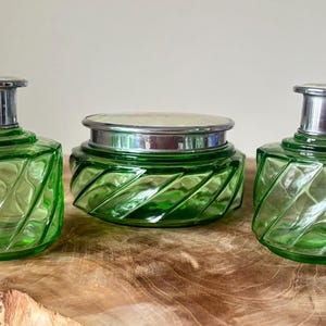 May include: A three-piece green glass vanity set with silver metal accents. The set comprises two bottles with stoppers and a jar with a lid. The glass features a diagonal, ribbed design. The set is displayed on a wooden surface.