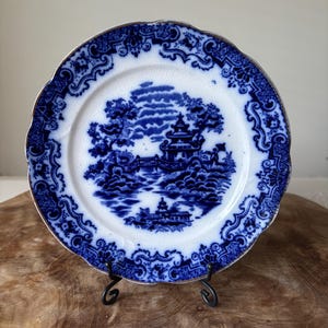 Puede incluir: Un plato decorativo blanco y azul con borde festoneado y ribete dorado. El plato presenta una escena de paisaje azul con una pagoda, árboles y un puente. El plato se exhibe sobre un soporte de metal negro.