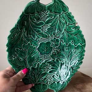 May include: A green ceramic leaf-shaped serving platter with intricate leaf detailing. The platter has a handle at the top and bottom, and is held by a hand with pink nail polish. The ceramic dish is a decorative piece for serving food.