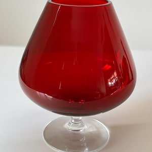 Könnte beinhalten: Ein großes, rubinrotes Brandyglas mit breitem Kelch und klarer, kreisförmiger Basis. Das Glas hat eine glänzende Oberfläche und reflektiert das Licht. Der Stiel ist kurz und verbindet den Kelch mit dem Fuß. Die Form ist abgerundet und elegant.