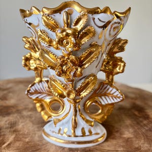 May include: A white and gold ceramic vase with a floral design. The vase features gold-colored roses and leaves, with gold trim along the edges. The vase is approximately 6 inches tall.