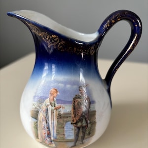May include: A small white porcelain pitcher with a blue and gold design. The pitcher features a painted scene of a man and woman standing on a bridge.