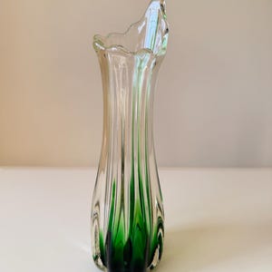 May include: A tall, slender glass vase featuring a clear upper section that transitions into a deep emerald green base. The vase has a vertical ribbed texture throughout its body and an asymmetrical, wavy, scalloped rim at the top opening.