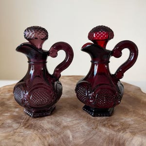 May include: Two small glass cruets with matching stoppers, one in a deep purple hue and the other in a vibrant ruby red. Each vessel features a textured hobnail pattern on the body, a curved handle, a flared spout, and a faceted, rounded stopper.