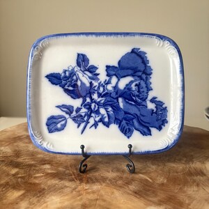 May include: Rectangular white ceramic dish with a blue floral design. The dish has a blue border and is supported by a black metal stand. The floral pattern features roses and leaves.