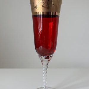 Può includere: Un elegante calice da champagne riempito con un liquido rosso intenso. Il bicchiere ha un gambo trasparente e attorcigliato e una fascia dorata sulla parte superiore con un motivo floreale. Il bicchiere è su una superficie bianca.