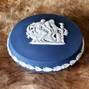 Puede incluir: Una caja de baratijas redonda azul con un diseño en relieve blanco en la tapa. El relieve representa figuras clásicas. La caja tiene un patrón de hojas blancas alrededor del borde. La caja está sobre una superficie de madera.