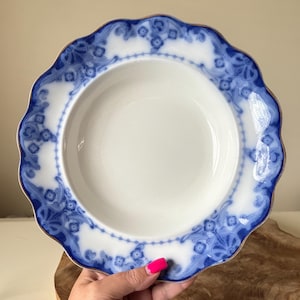 May include: White ceramic bowl with a blue floral design around the rim and a gold trim. The bowl has a scalloped edge and is held by a person with pink nail polish.