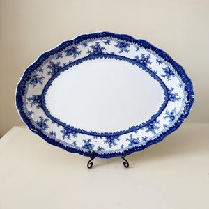 May include: An oval ceramic serving platter featuring a white centre and a wide blue and white border. The border displays a repeating geometric Greek key pattern along the inner edge and a detailed floral motif around the scalloped rim.