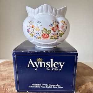 May include: A white Aynsley bone china vase with a scalloped rim, adorned with colorful floral patterns. The vase sits atop a navy blue box with the Aynsley logo and text: "Est. 1775". The box also states "Makers of the Finest English Bone China."