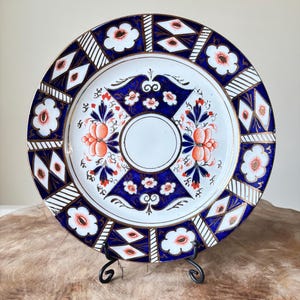 Puede incluir: Un plato decorativo con un centro blanco y un borde con diseños azules y blancos. El plato presenta diseños florales naranjas y ribetes dorados. Se exhibe sobre un soporte de metal negro.