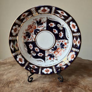 Puede incluir: Un plato decorativo de porcelana con un diseño floral y geométrico detallado. El plato presenta un centro blanco, cuadrantes azul marino y blancos y un borde dorado. Los elementos florales son en tonos naranja y blanco. El plato se exhibe sobre un soporte de metal negro.