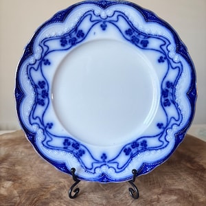 May include: A white and blue decorative plate with a scalloped edge. The plate has a blue floral design around the rim and a white centre. It is displayed on a black metal stand, highlighting its intricate details.