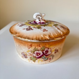 Puede incluir: Una caja de porcelana vintage con un diseño floral. La tapa tiene un pequeño pomo en forma de pájaro blanco. La caja está decorada con un borde dorado y un patrón floral en tonos de morado, amarillo y verde.