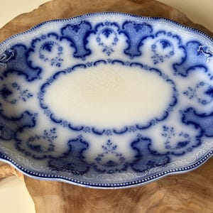 May include: An oval-shaped, antique serving platter with a white center and a detailed blue floral pattern around the edges. The platter has a scalloped rim and is placed on a wooden surface.