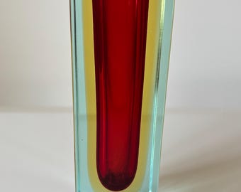 May include: A tall, rectangular glass vase featuring a layered design with a deep red cylindrical center, a yellow middle layer, and a clear light blue outer casing. The vessel has a flat base and a wide, open top with thick, smooth glass walls.