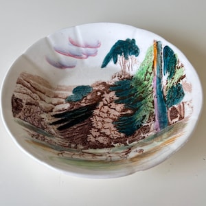 May include: A white ceramic plate with a landscape design. The plate features a painted scene with trees, a waterfall, and a mountain range in shades of green, brown, and blue. The rim of the plate has a scalloped edge.
