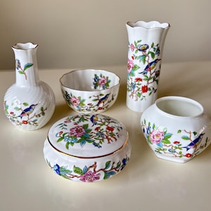 May include: A collection of white ceramic decorative pieces with floral and bird designs. The set includes a vase, a bowl, a lidded box, and a small vase. The pieces feature a gold trim and are decorated with colorful flowers and birds.