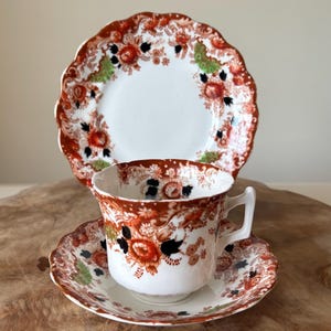 May include: A vintage teacup, saucer, and plate set. The white porcelain features a floral pattern in shades of red, brown, black, and green. The edges of the plate and saucer have a scalloped design. The teacup has a handle.