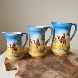 May include: A set of three ceramic pitchers in graduated sizes, each featuring a painted desert scene with a person riding a camel, palm trees, and pyramids against a blue sky and yellow sand background, with a curved handle and pouring spout.