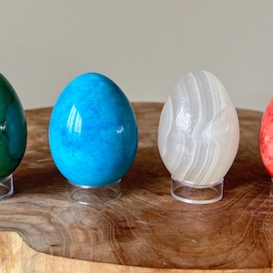 Puede incluir: Cuatro huevos de piedra pulida en tonos verde, azul, blanco y rojo, cada uno sobre un soporte de acrílico transparente. Los huevos se exhiben sobre una superficie de madera natural, mostrando sus superficies lisas y reflectantes y sus variados colores.