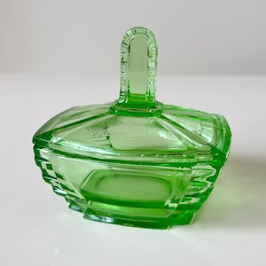 Könnte beinhalten: Eine lichtdurchlässige, hellgrüne Glasschale mit passendem Deckel. Die Schale hat eine rechteckige Form mit abgestuften Seiten und einen zentralen vertikalen Griff auf dem Deckel, der durchgehend mit einem gekerbten Kantendesign versehen ist.