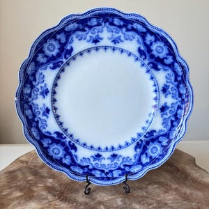 May include: A white and blue ceramic plate with a scalloped edge. The plate features a detailed blue floral pattern around the rim and a smaller floral design near the center. The plate is displayed on a black metal stand.