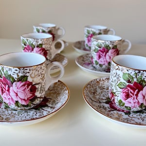 Könnte beinhalten: Sechs Vintage-Tassen und Untertassen. Die weißen Tassen und Untertassen sind mit einem goldenen Filigranmuster und großen rosa Rosen verziert. Die Ränder sind goldfarben.
