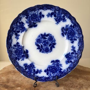 May include: A white and blue antique plate with a floral design. The plate has a scalloped edge and is supported by a black metal stand. The blue floral pattern is repeated around the rim and in the center of the plate.