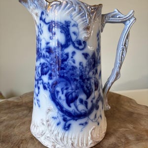 Puede incluir: Jarra vintage blanca y azul con diseño floral. La jarra tiene un asa detallada y un ribete dorado alrededor de la parte superior e inferior. El patrón floral azul es intrincado y cubre la mayor parte de la superficie de la jarra.