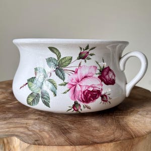 Könnte beinhalten: Weißer Keramiknapf mit Henkel, verziert mit einem floralen Rosendesign in Rosa, Rot und Grün. Die Schüssel hat eine breite, geschwungene Form und einen einzelnen Henkel. Das florale Muster ist detailliert und bedeckt einen Großteil der Oberfläche.