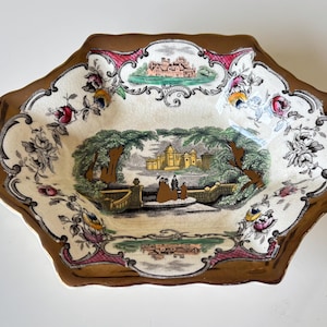 May include: An antique, octagonal serving dish with a brown rim and a cream-colored interior. The dish features a central landscape scene with figures and buildings, surrounded by floral and architectural motifs.