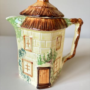 Può includere: Una teiera in ceramica a forma di cottage con tetto di paglia. La teiera è dipinta in tonalità di giallo, marrone e verde, con dettagli di finestre, una porta ed edera.